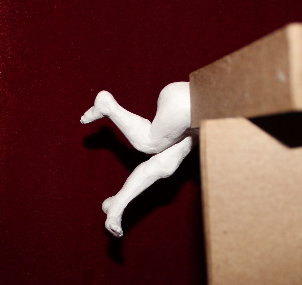 A pair of legs, made from clay, flail form a figure diving headfirst into a box.