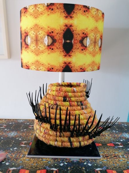 Coil basket using yellow fabric, with black spikes protruding in a spiral around the basket. Yellow lampshade to rear uses the same fabric print as the basket.